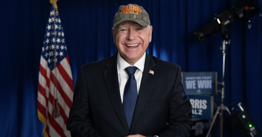 Tim Walz in a suit, wearing a camo-printed hat that says HARRIS WALZ in bright orange letters.